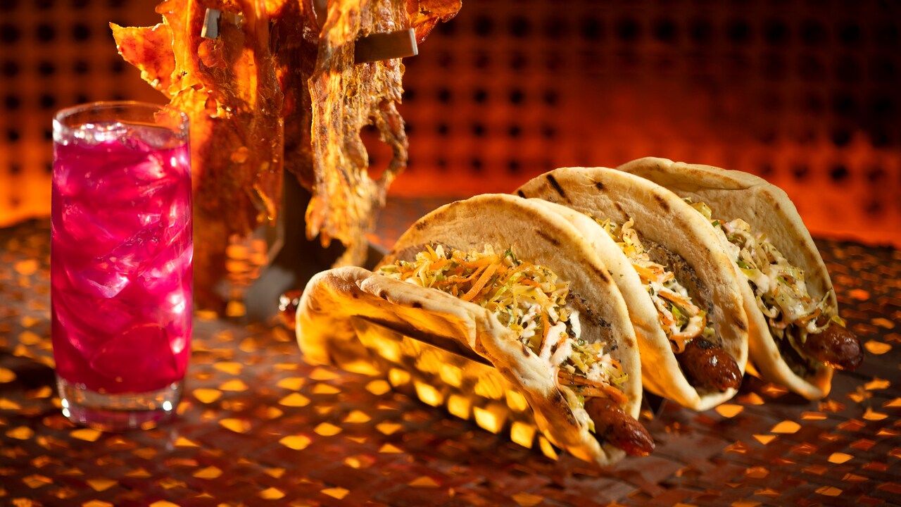 An exotic beverage and 3 thick tortillas filled with sausage, coleslaw and sauce