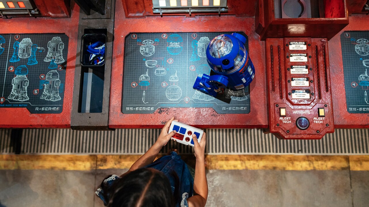 A young girl controls her new R series droid atop a workshop console featuring construction blueprints of droids.