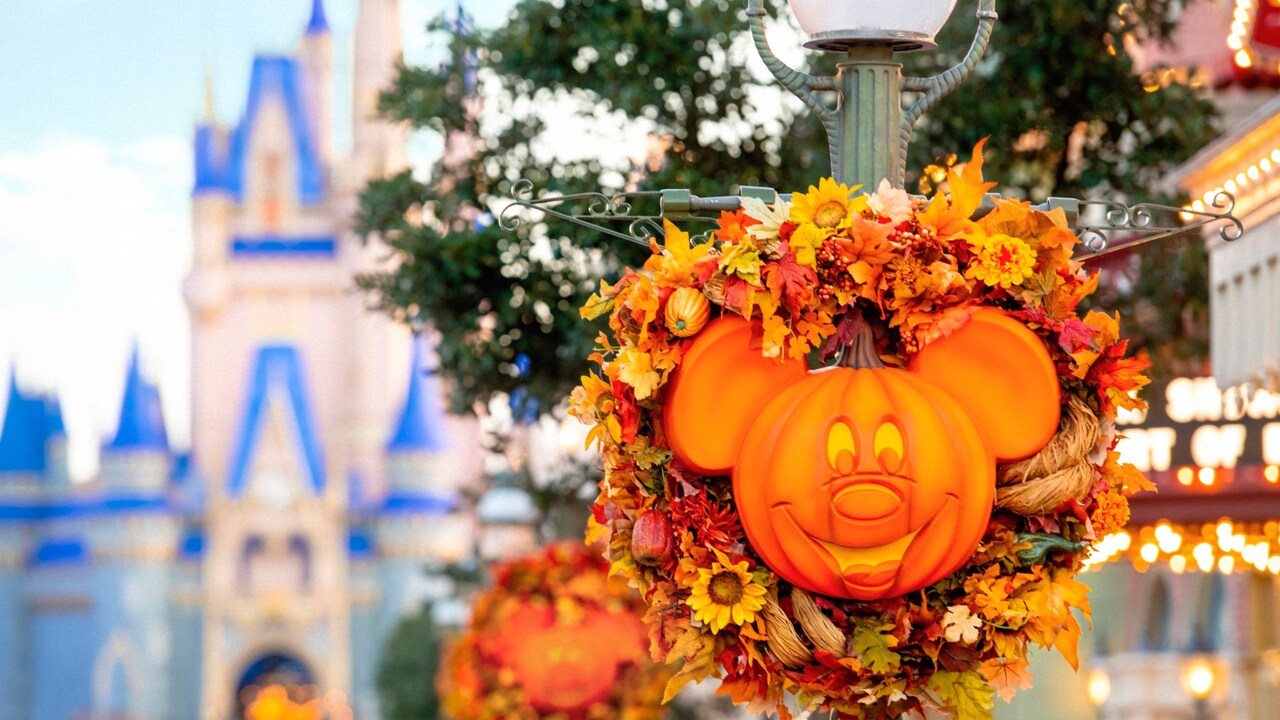 La parte superior de una lámpara de calle adornada con una corona temática de otoño rodea la cara y las orejas de una calabaza de Mickey Mouse con la frase ‘Autumn at Walt Disney World Resort’