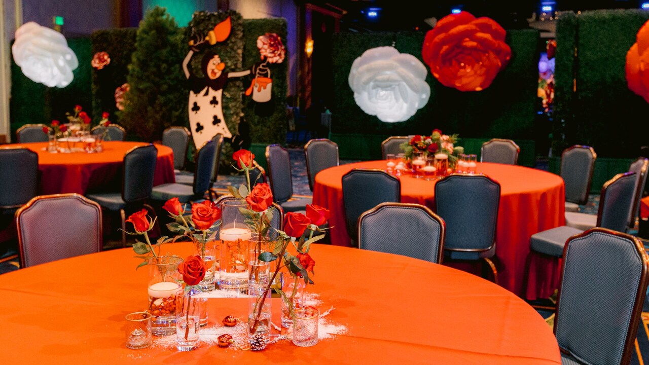Un espacio para eventos con salón de baile con mesas, sillas, arreglos florales y decoraciones que recrean el jardín de rosas de Queen of Hearts de Alice in Wonderland
