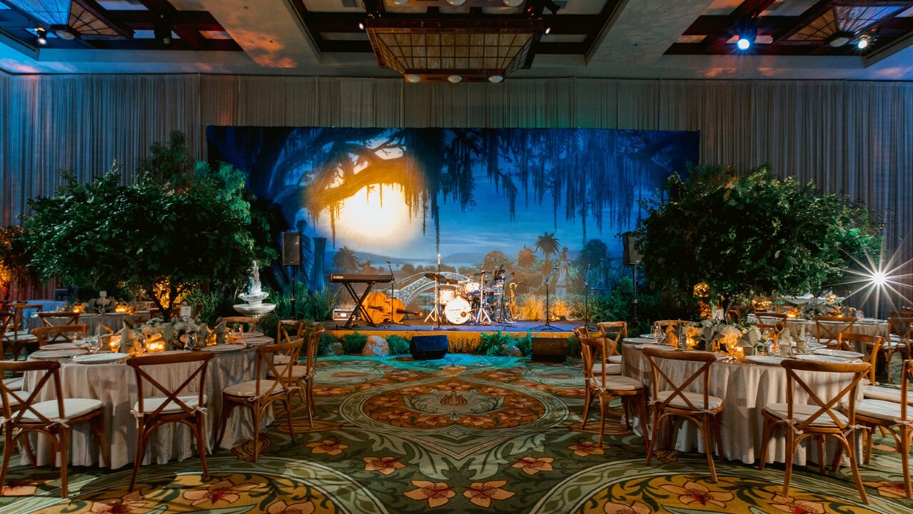 Un lugar para eventos con salón de baile con mesas de comedor, sillas, arreglos florales y un escenario con instrumentos musicales y un fondo de pantanos flanqueado por árboles