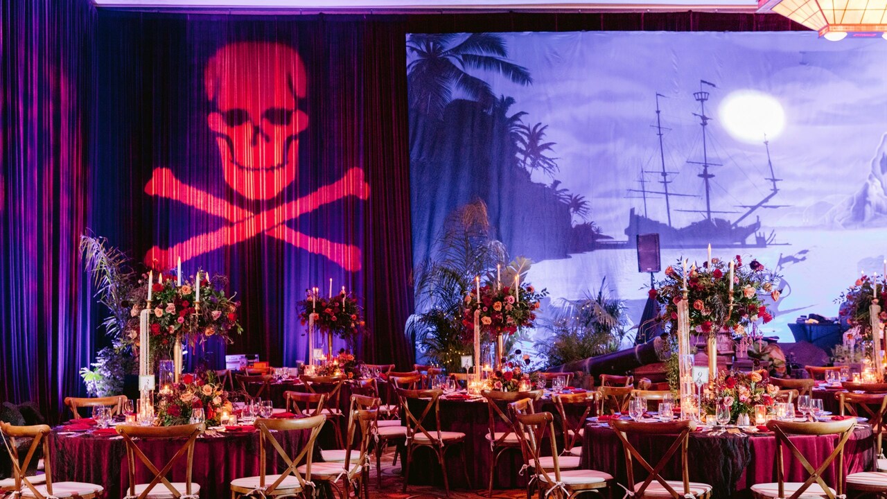 Un lugar para eventos con salón de baile con mesas de comedor, sillas, velas, arreglos florales y un telón de fondo con un barco pirata en una bahía y una proyección de una calavera y huesos cruzados