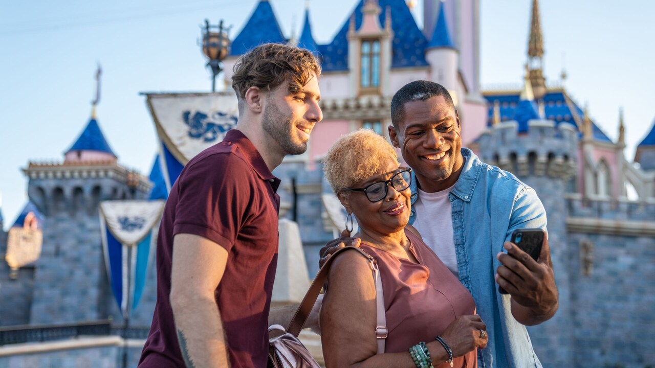 Dos hombres y una mujer miran un teléfono inteligente cerca del Sleeping Beauty Castle en Disneyland Park