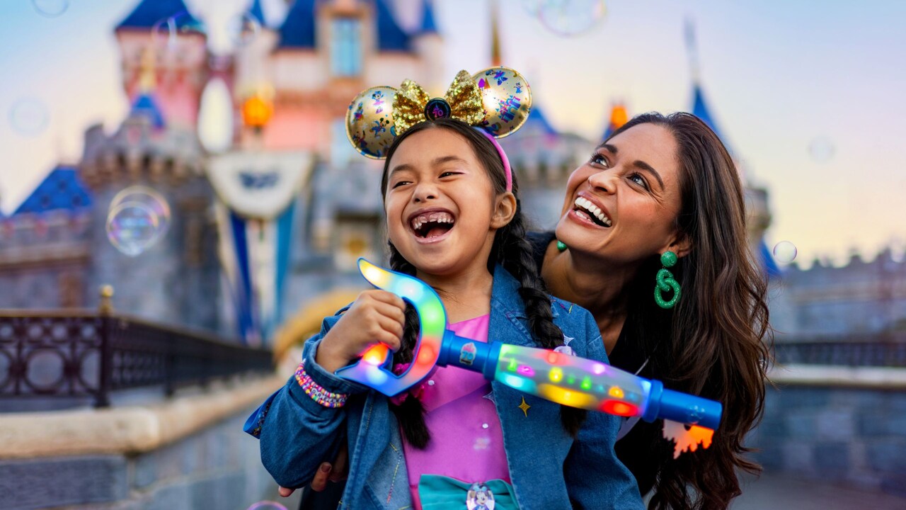 A girl wearing a Disneyland 70th anniversary ear headband and her mother, both playing with a bubble blower near Sleeping Beauty Castle