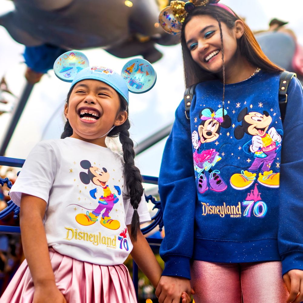 Two Guests wearing Disneyland Resort 70th Celebration apparel, including a Mickey ear hat, T-shirt and sweatshirt standing in front of the Dumbo the Flying Elephant attraction