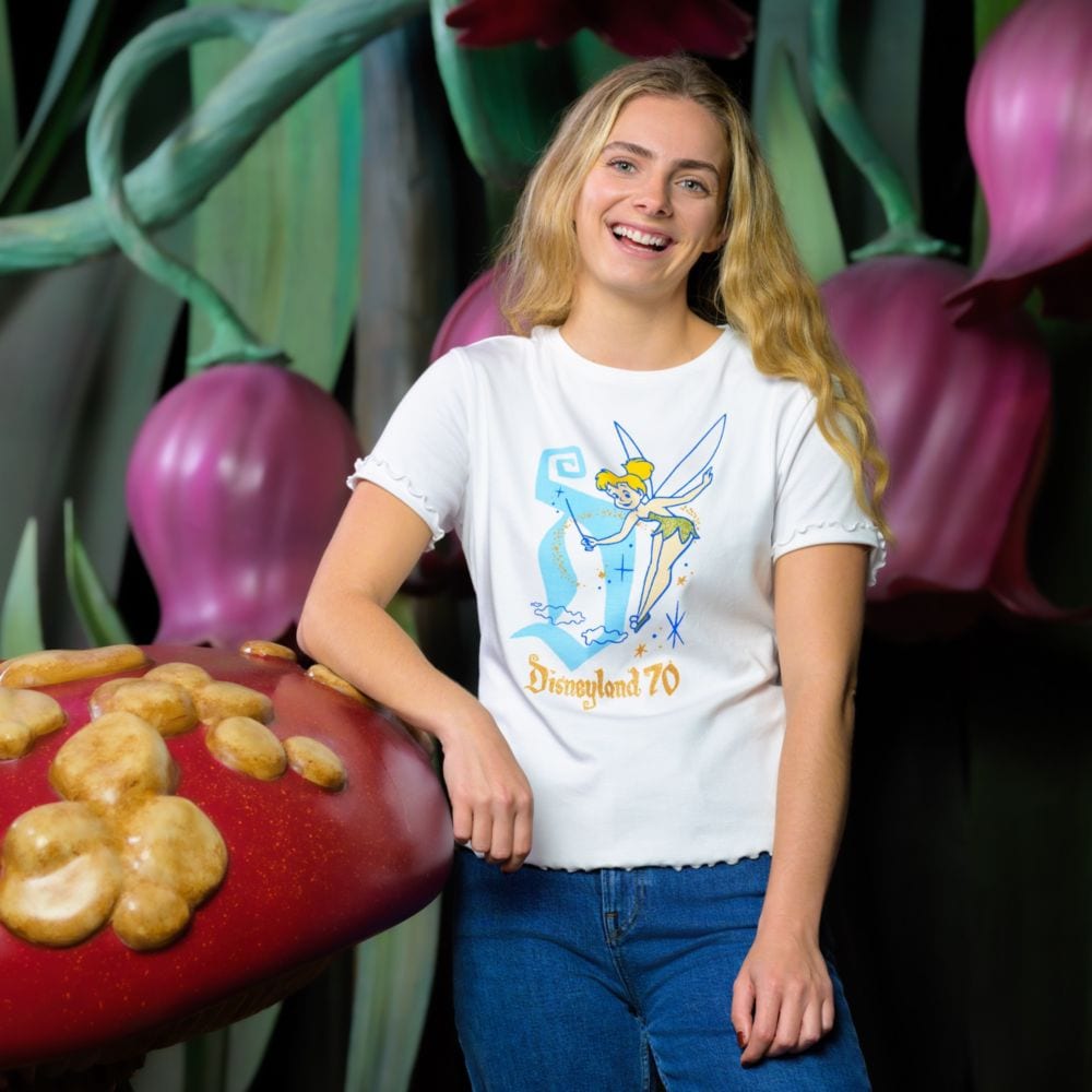 A woman smiling while wearing a shirt from the Disneyland 70th Anniversary Tinker Bell Collection as she leans on a giant mushroom