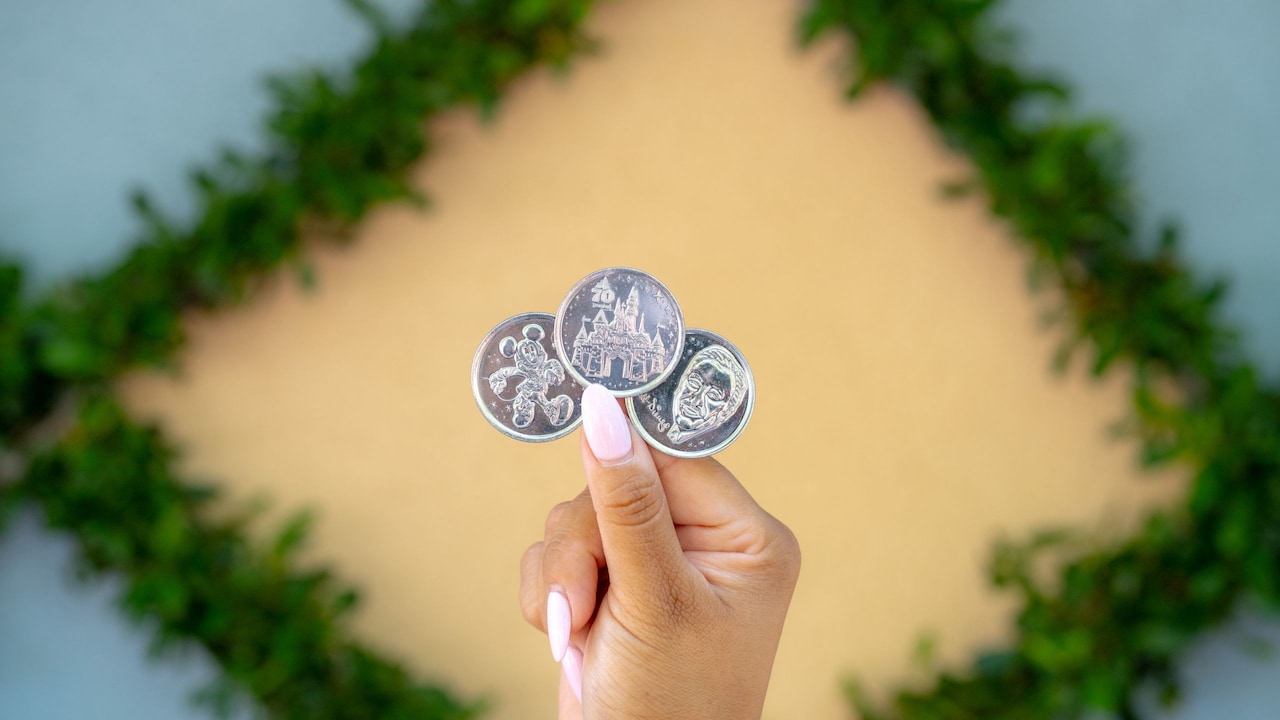 A hand holding 3 collectible Disneyland 70th Celebration medallions featuring Mickey Mouse, Sleeping Beauty Castle and Walt Disney