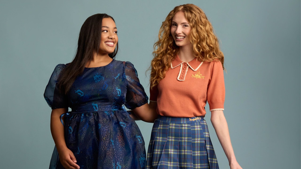 Two women, one wearing a dress featuring a print with Sleeping Beauty Castles and the Disneyland ‘D’ logo and the other wearing a knit blouse and a skirt featuring embroidered text that says ‘Disneyland 70’