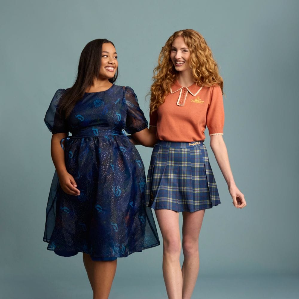 Two women, one wearing a dress featuring a print with Sleeping Beauty Castles and the Disneyland ‘D’ logo and the other wearing a knit blouse and a skirt featuring embroidered text that says ‘Disneyland 70’