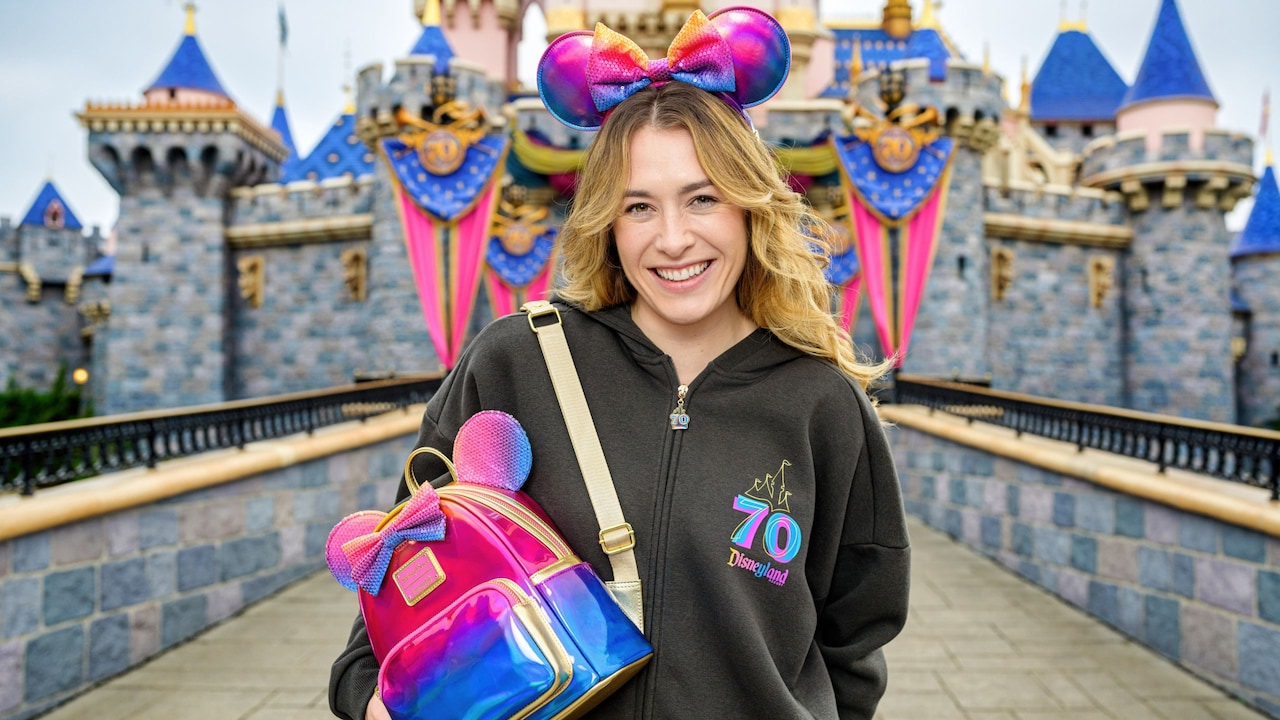 A Guest wearing a hoodie and Minnie Mouse ear headband, holding a lounge fly backpack from the Disneyland Resort 70th Celebration "Celebrate Happy Collection" in front of Sleeping Beauty Castle at Disneyland park