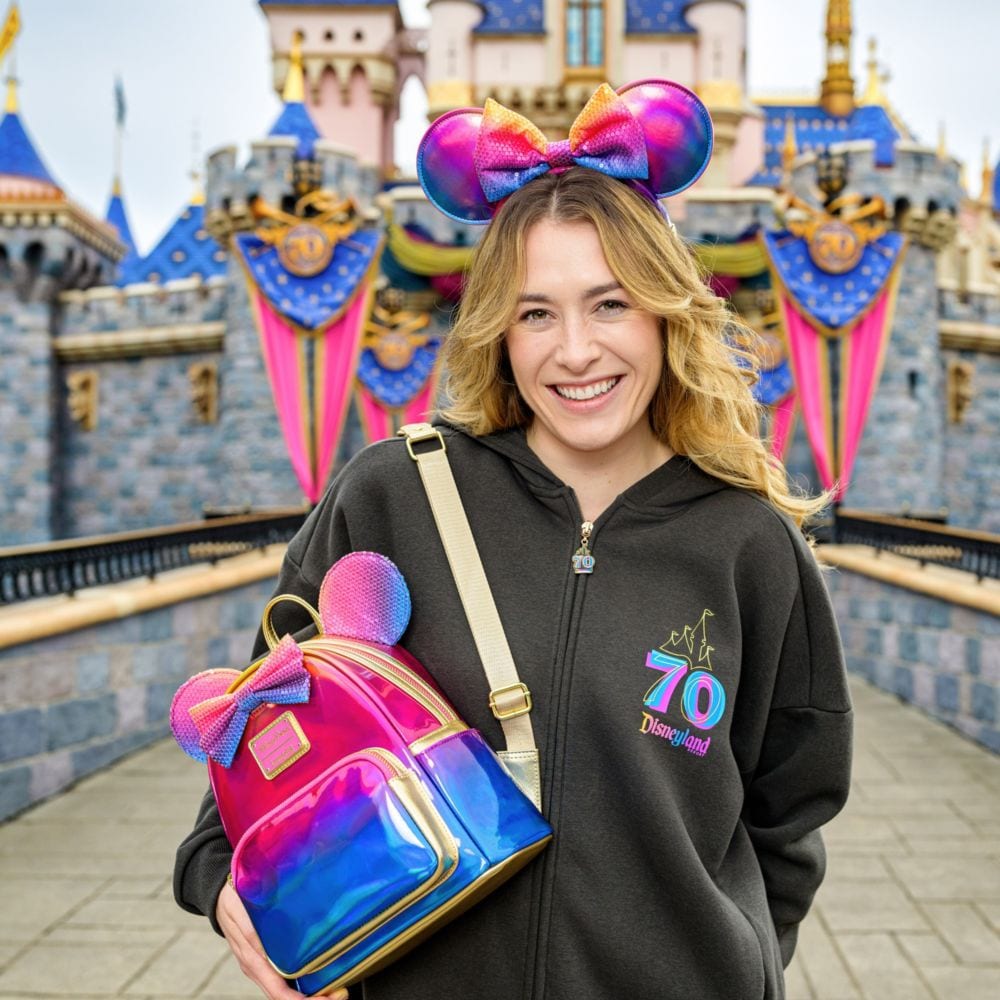 A Guest wearing a hoodie and Minnie Mouse ear headband, holding a lounge fly backpack from the Disneyland Resort 70th Celebration ‘Celebrate Happy Collection’ in front of Sleeping Beauty Castle at Disneyland park