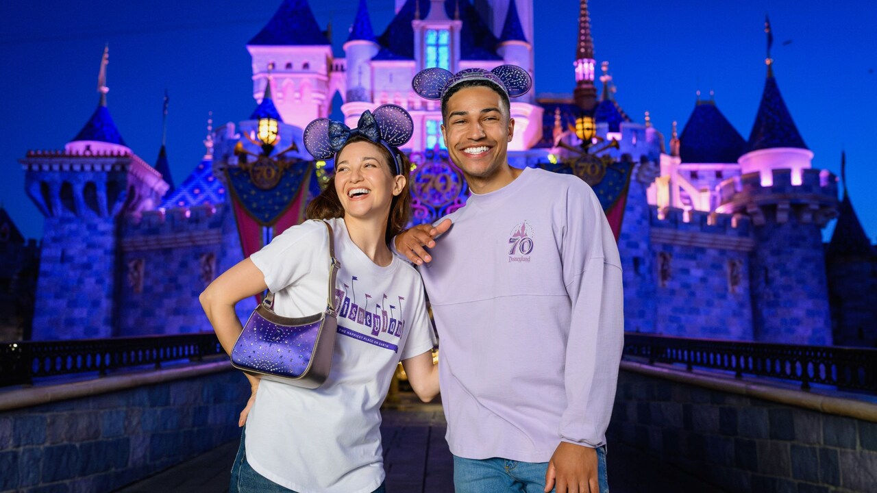 Two Guests standing near Sleeping Beauty Castle wearing merchandise commemorating the Disneyland 70th Celebration