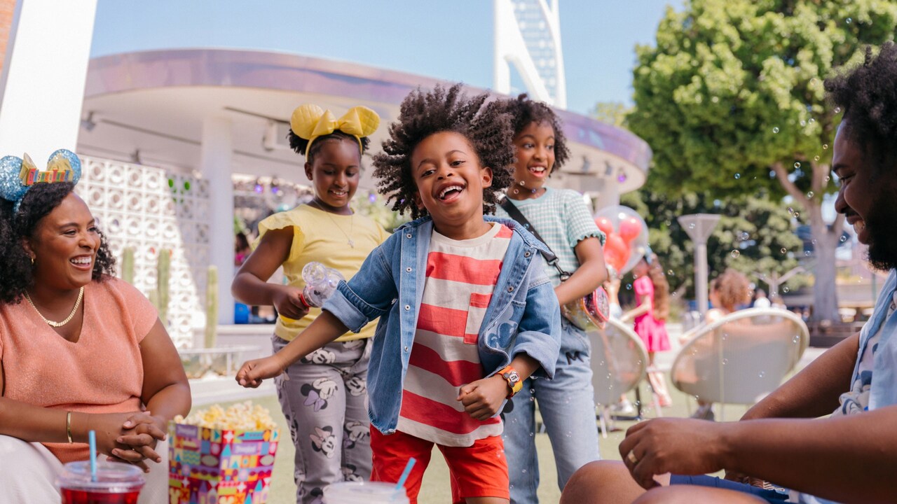 Three children dance and play as 2 adult family members look on, next to the Downtown Disney Live Stage