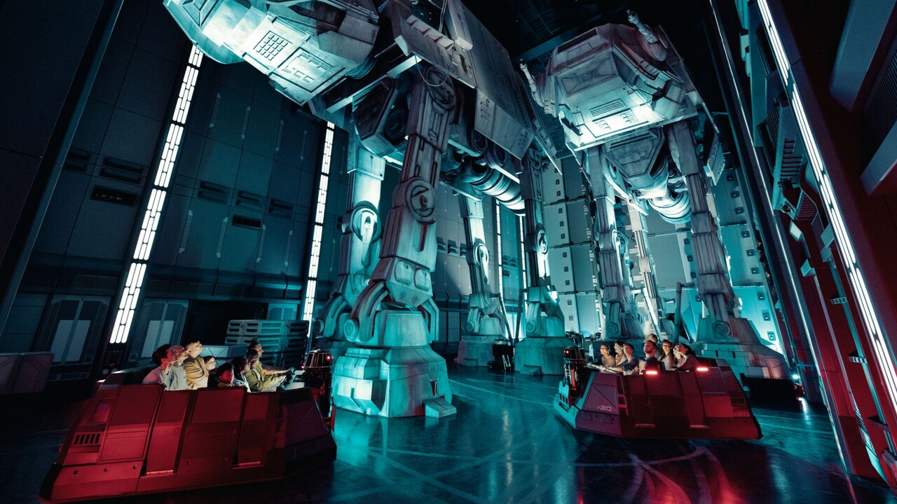  People in a transport vehicle inside a hanger bay look up at 2 towering AT-AT walkers