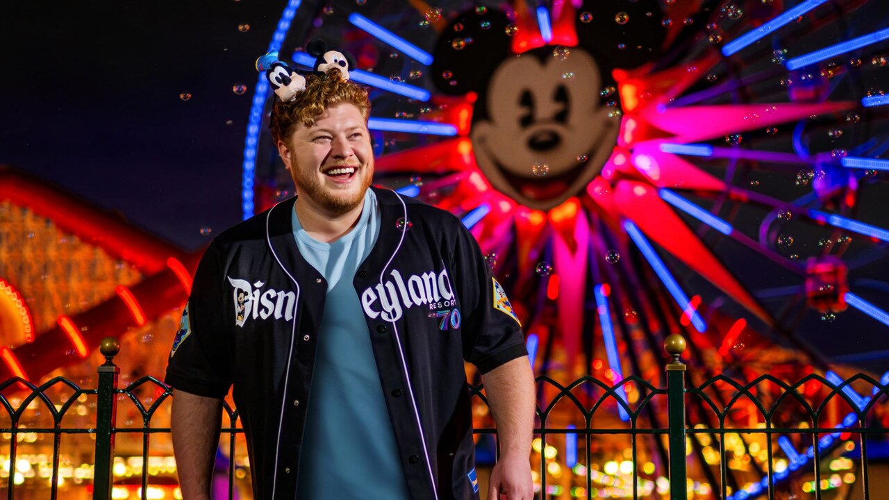 Visitante con una camiseta de béisbol del 70.º Aniversario de Disneyland y una diadema con orejas de Mickey Mouse, de pie junto a una barandilla con una noria iluminada que tiene el rostro de Mickey detrás, por la noche