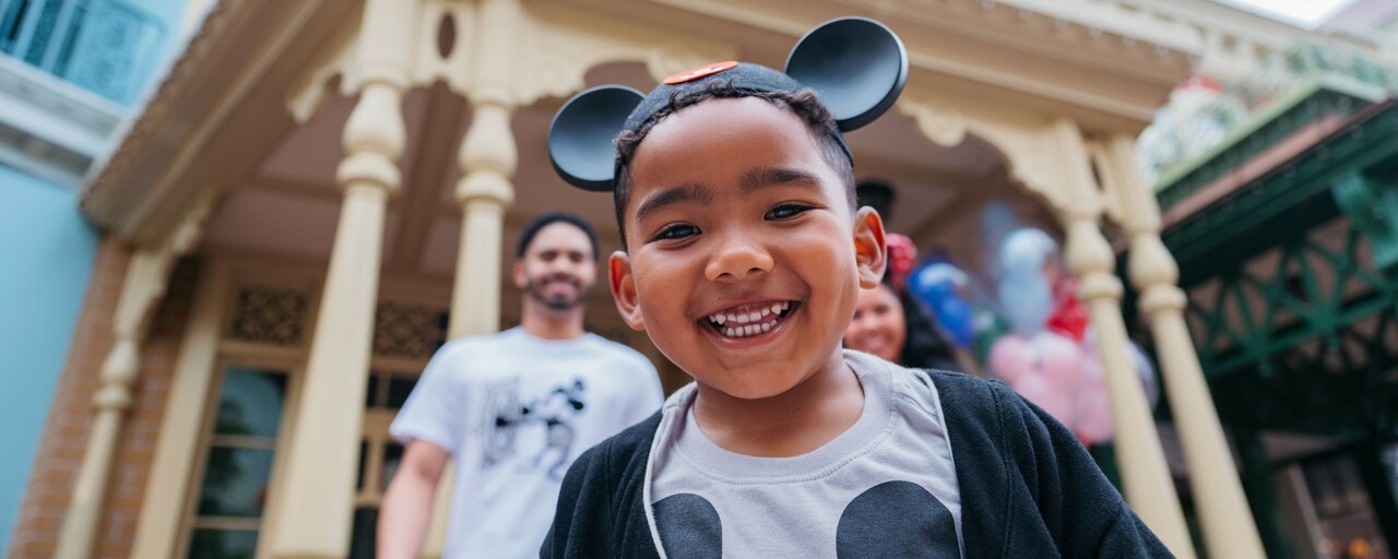 Un niño sonriente de pie frente a sus padres con un sombrero con orejas y una camisa de Mickey Mouse