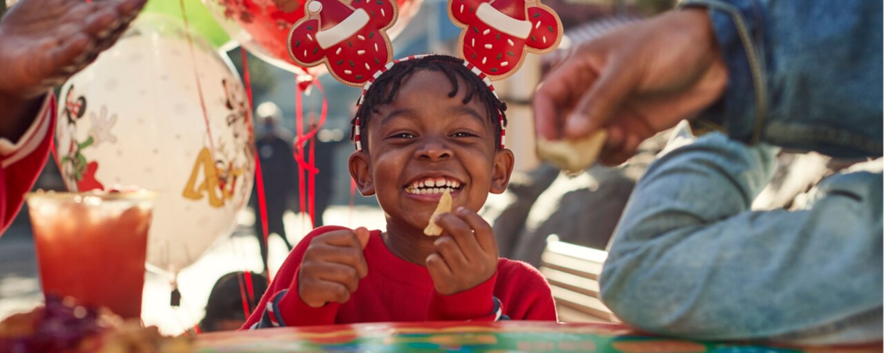 A smiling young girl in a Santa themed Minnie bow eating a snack chip