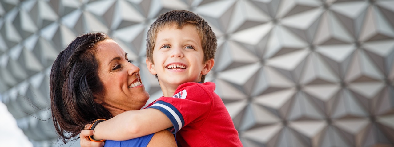 A smiling mother holds her young son in his arms in front of Spaceship Earth at EPCOT 