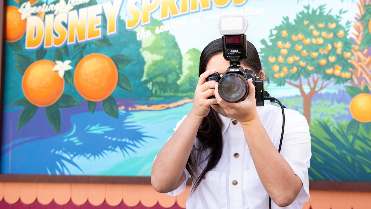 A Disney PhotoPass photographer points her camera in front of a sign that says ‘Greetings from Disney Springs’