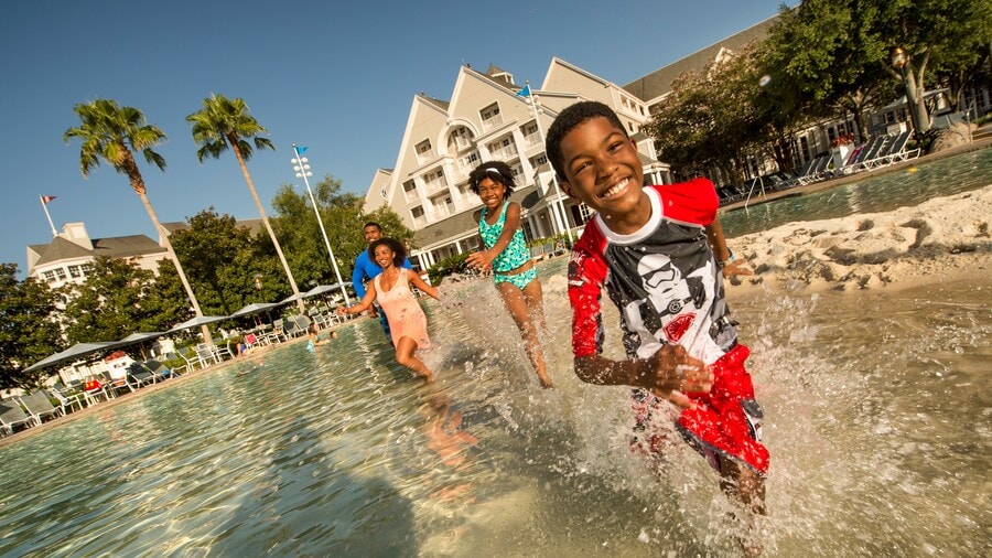 A smiling boy splashes through knee high water with his family watching in the background