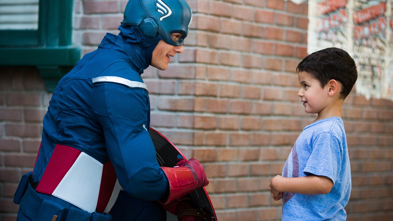 El Capitán América, con su escudo a un lado, se hinca para saludar a un niño