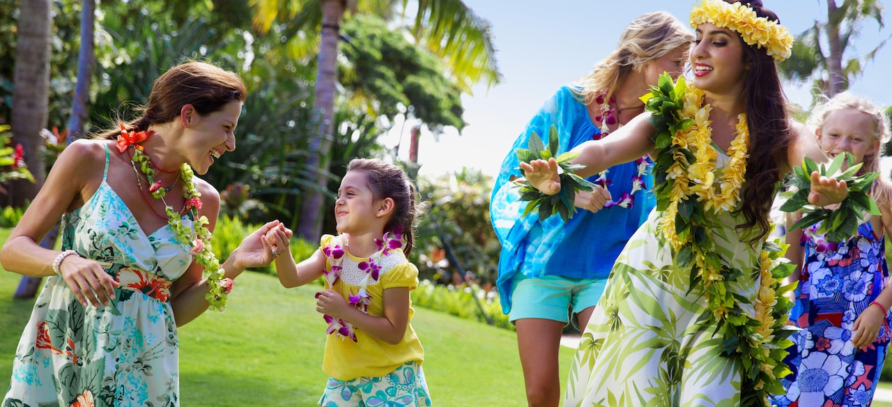 Two young girls and their mothers wearing flower leis follow along during a Hawaiian hula dancing lesson