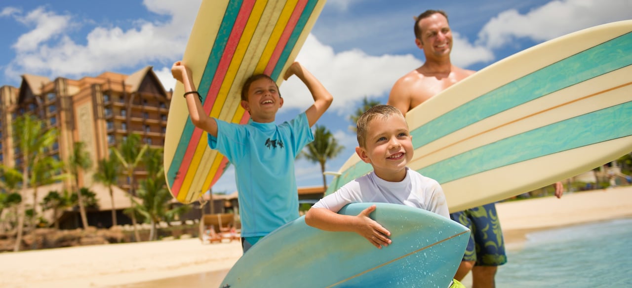 A father and his 2 young sons head toward the ocean carrying surfboards