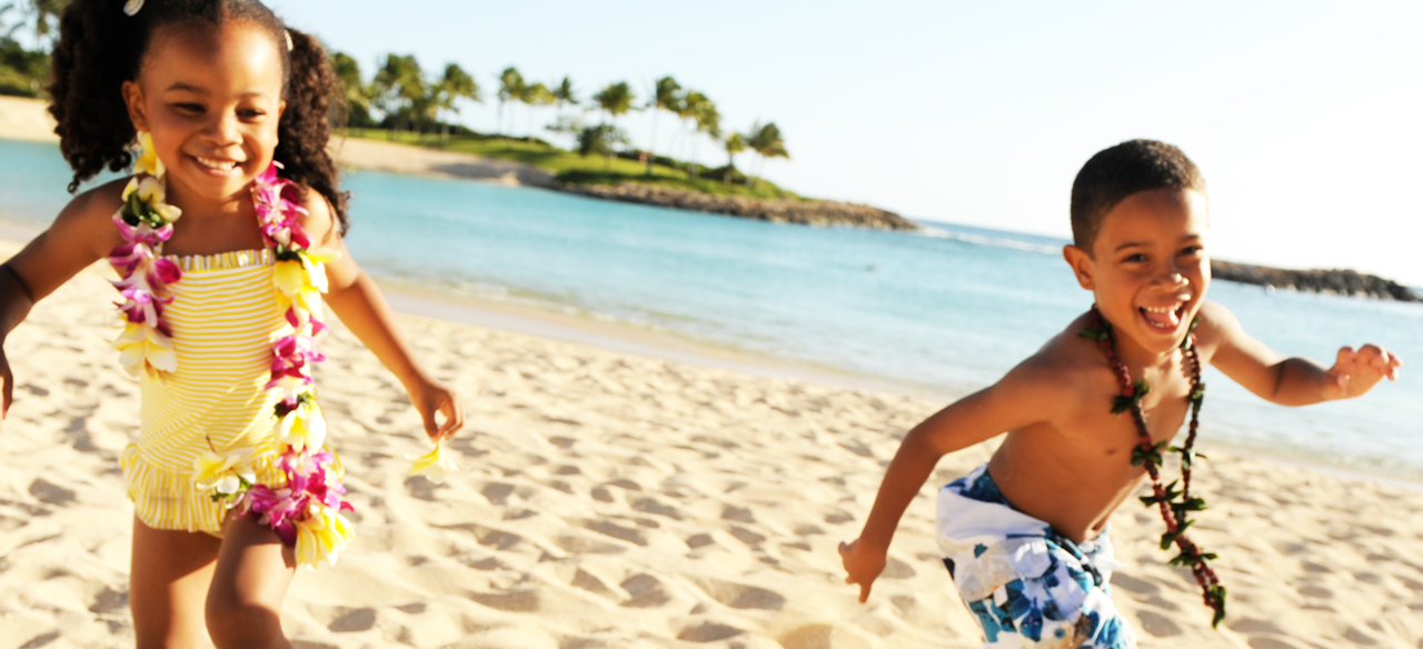 A small girl and boy wearing swimwear and Hawaiian necklaces frolic on a tropical beach