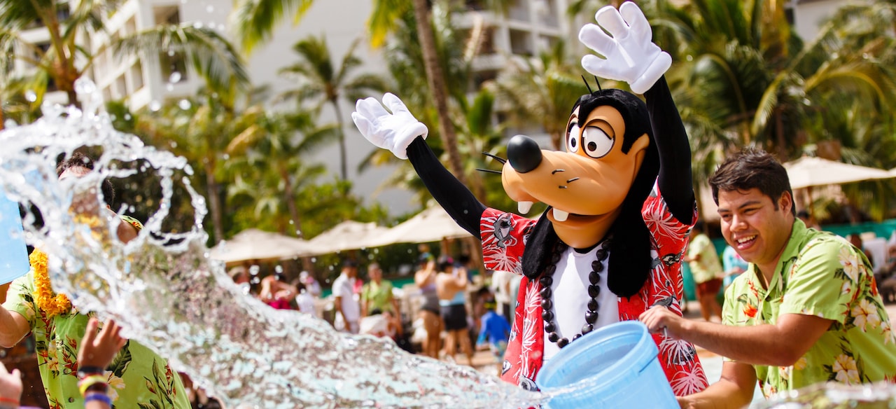 A Cast Member throws a bucket of water while Goofy, dressed in Hawaiian garb, waves his hands in the air