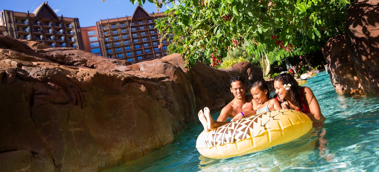 A girl on an inner tube in a lazy river with her father and mother, with rocky banks and a tree overhead