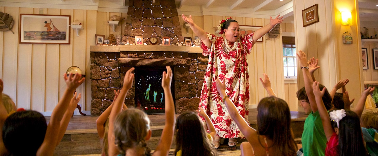 A woman in Hawaiian garb and a group of children sit on the floor watching her reach to the sky