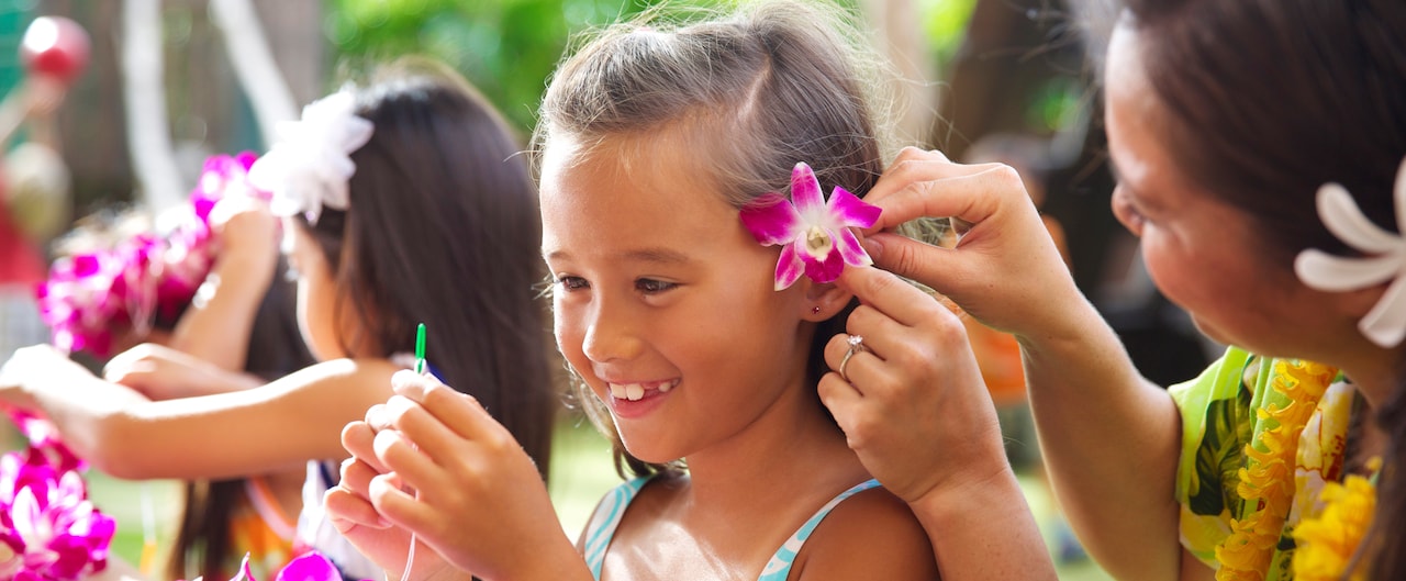 A Cast Member tucks a fuchsia orchid behind a little girl's ear as she threads flowers to make a lei