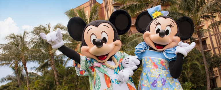 Mickey Mouse and Minnie Mouse, dressed in Hawaiian style outfits, stand in front of a wing of Aulani Resort