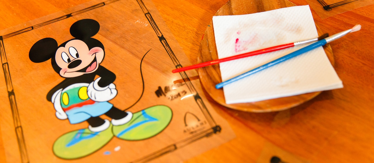 An illustration of Mickey Mouse on a wood surface adjacent to a dish with paintbrushes resting on a napkin