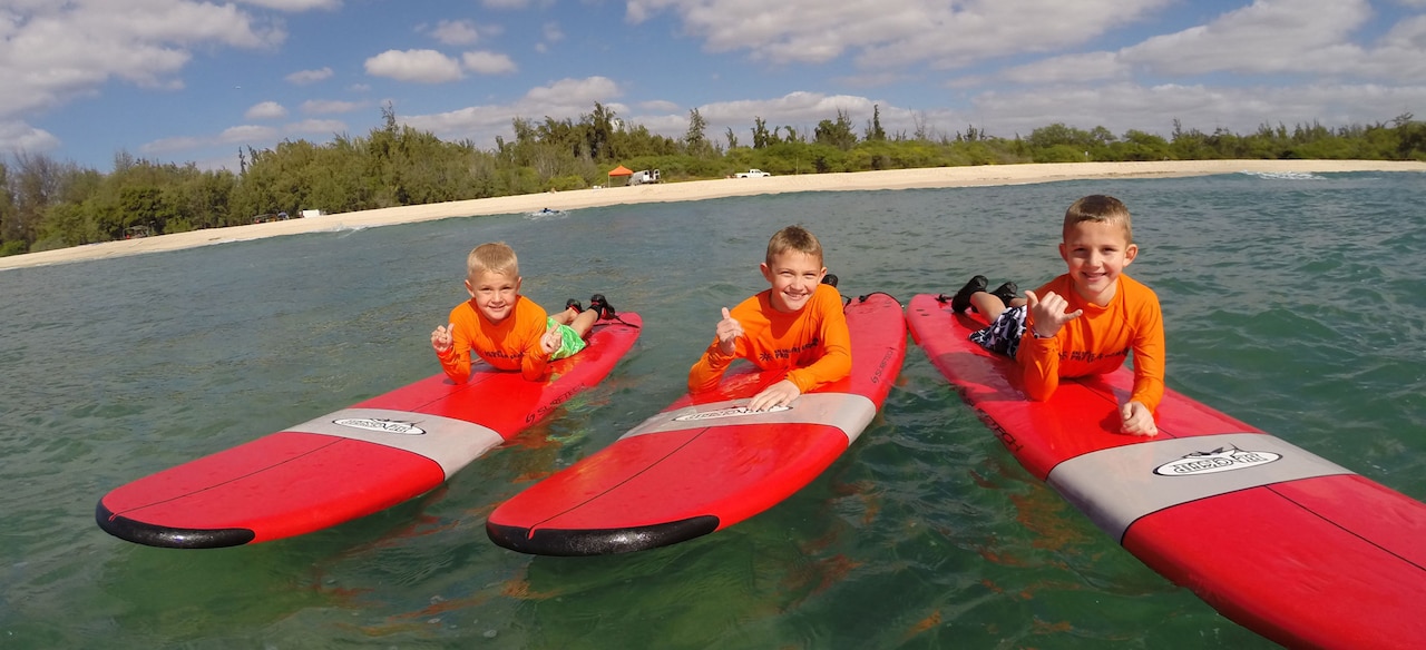 Three young boys give the hang loose hand sign as they lie on their bellies on surfboards floating in the ocean