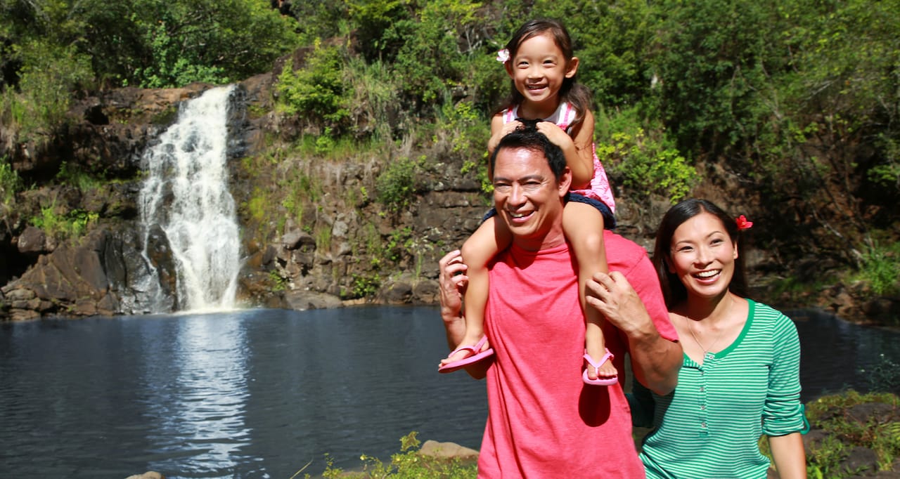 A woman and a man with a young girl on his shoulders walk beside a pond near a waterfall