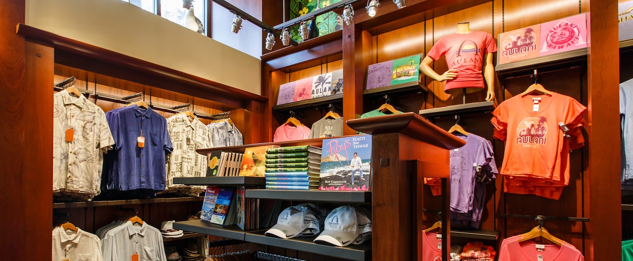 Aulani caps and books about Hawaii on a shelf, with men's shirts and tees hanging nearby