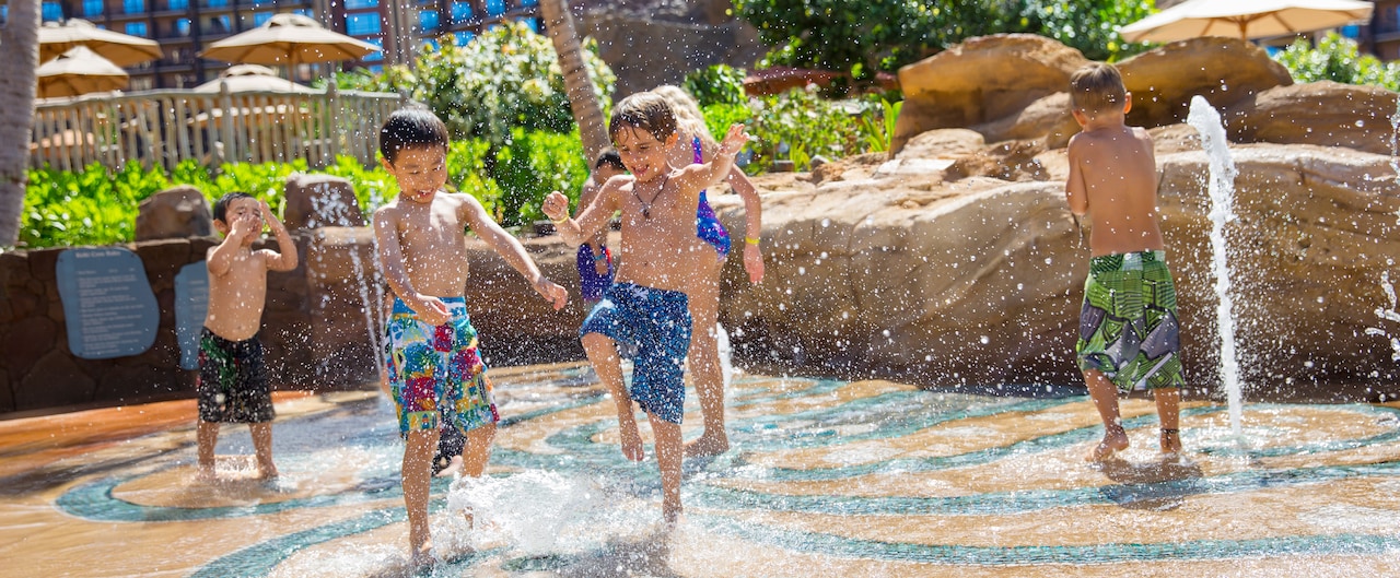 Two small boys in swim shorts play around squirting water jets near 4 more playing children