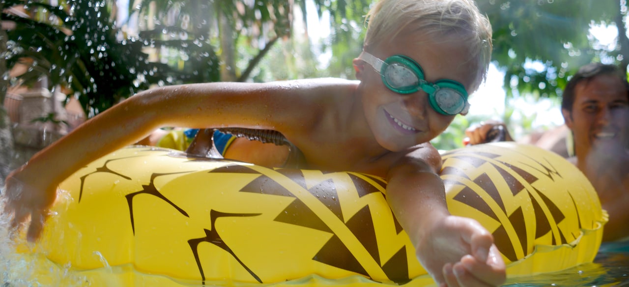 A smiling boy wearing swim goggles lies on top of a yellow inner tube on a lazy river