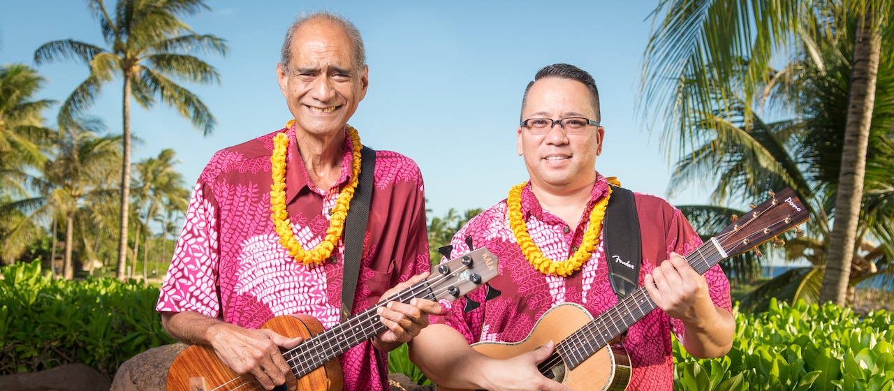 Musician and storyteller Hoku Zuttermeister, in Aloha shirt and lei, holds a ukulele and stands beside a man holding a guitar wearing a matching shirt and lei