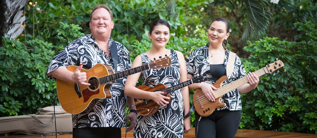 The musical trio Kapena, one man holding a guitar and two women, both holding their instruments, stand amid the Aulani foliage wearing matching Hawaiian print outfits