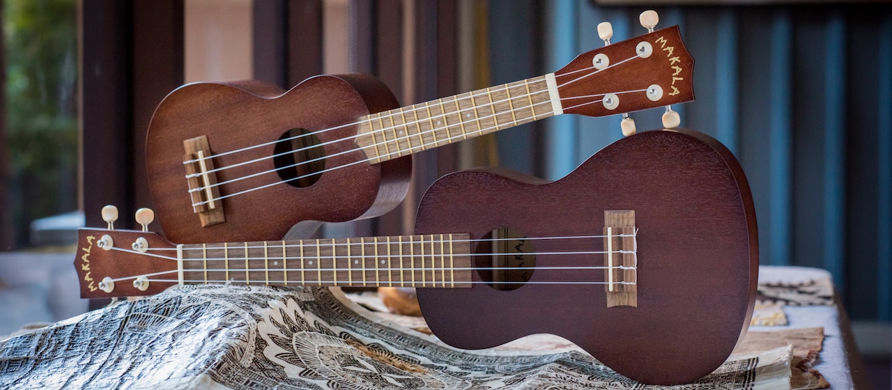 Two &#8216;ukuleles, one on top of the other, lay on their sides on a table with a small mat beneath them
