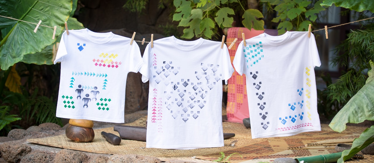 Three t-shirts with kapa stamp designs are hung on a line with clothespins