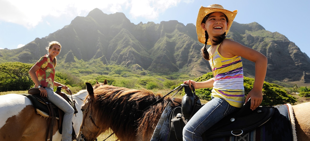 Two teen girls ride horses along a grassy plain with mossy mountains looming beyond