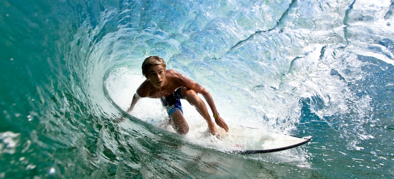 A teen boy rides a surfboard inside the barrel of a large wave