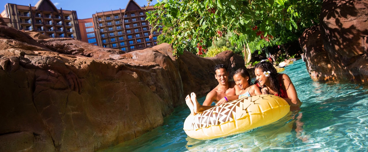 A girl on an inner tube near her father and mother along a lazy river, with rocky banks and a tree
