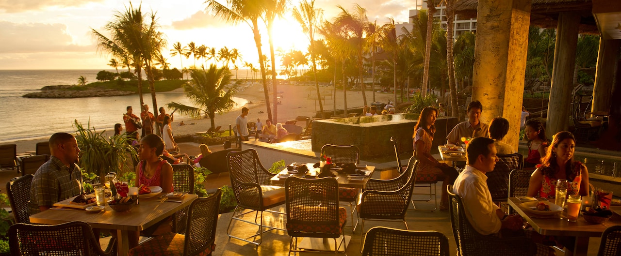 Two couples and a party of 4 dine on an outdoor patio surrounded by palm trees and ocean at sunset