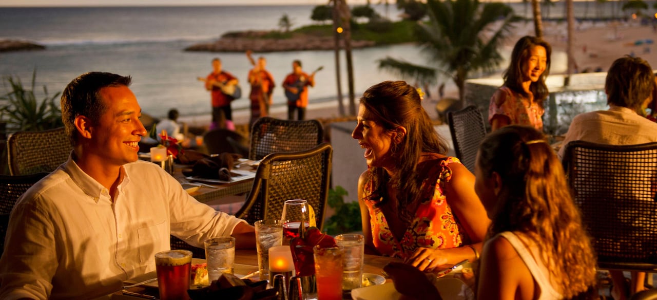 A father, mother and daughter enjoy drinks at an outdoor table with sunset ocean views and live musicians