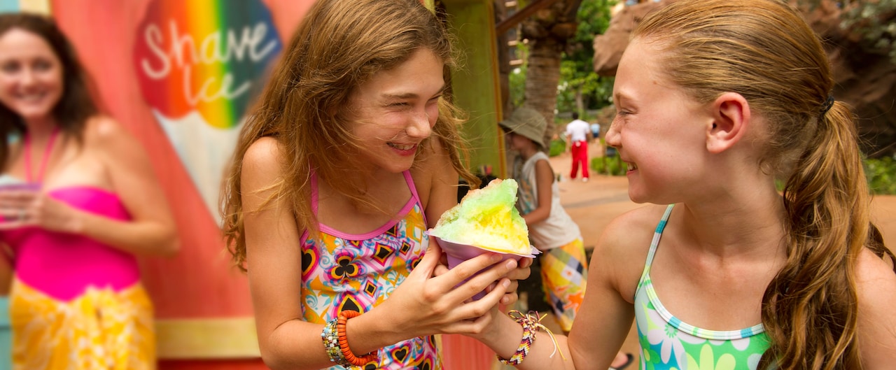 Two tween girls in swimwear laugh while clutching a shave ice dessert as a woman holding a shave ice looks on