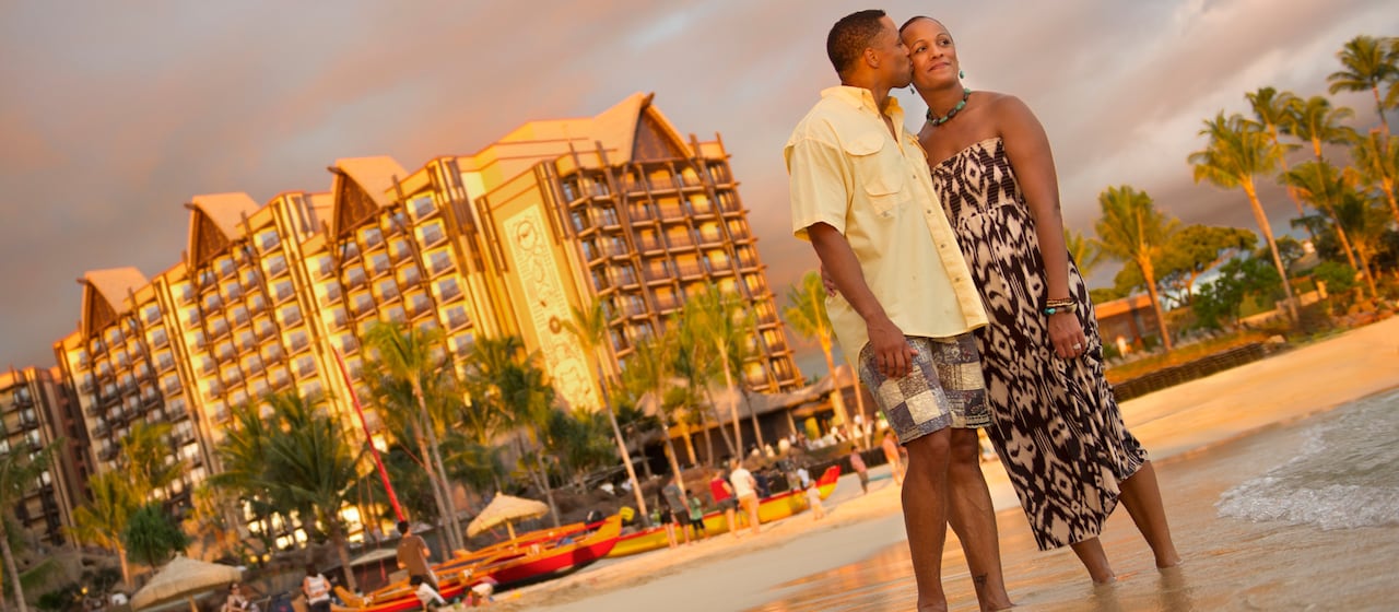 A man kisses a woman on the cheek as they stand on the shore in front of Aulani Resort